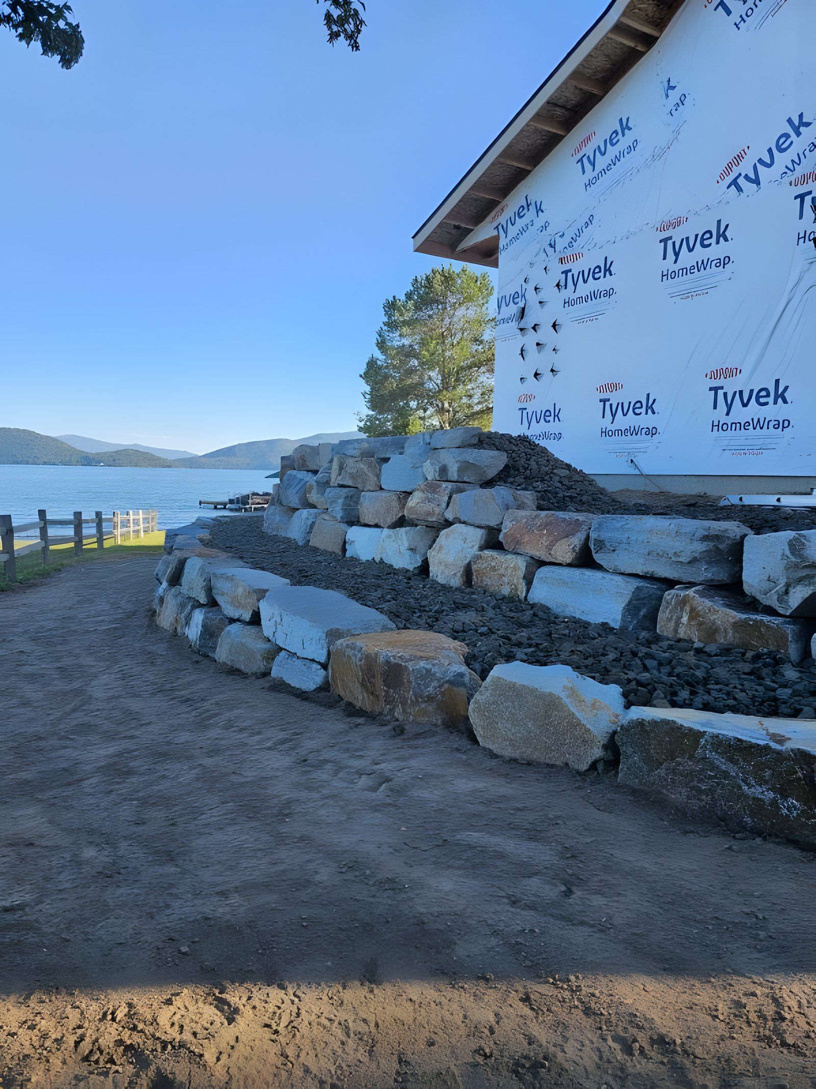 Two-tier boulder retaining wall in front of a house in Tyvek wrap, with the lake and mountains behind it