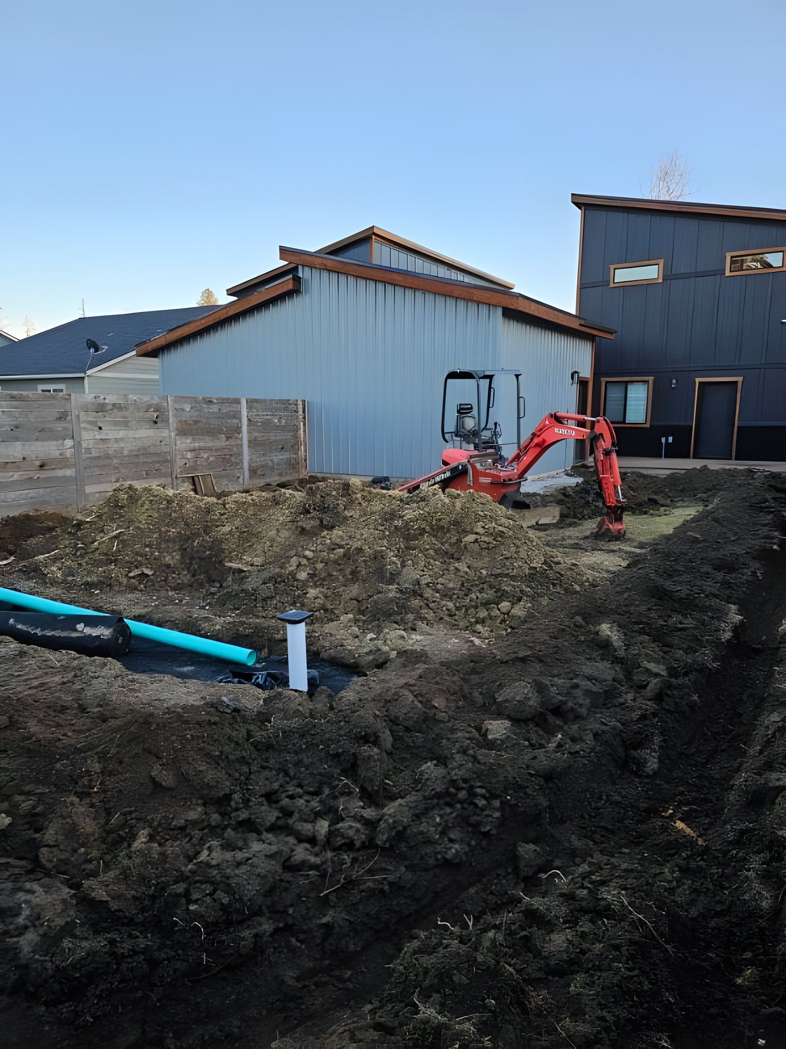 Trench in a backyard with green drain pipe and a mini excavator next to a new house