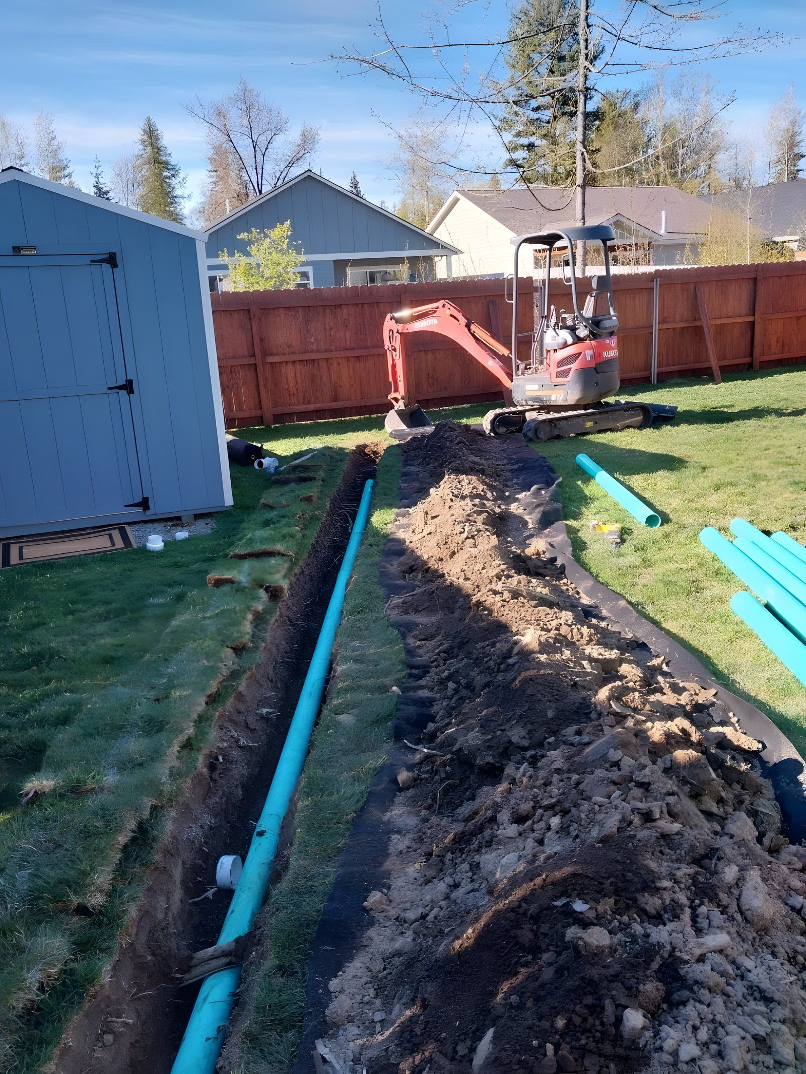 Trench through a backyard lawn with green pipe laid alongside and a mini excavator behind